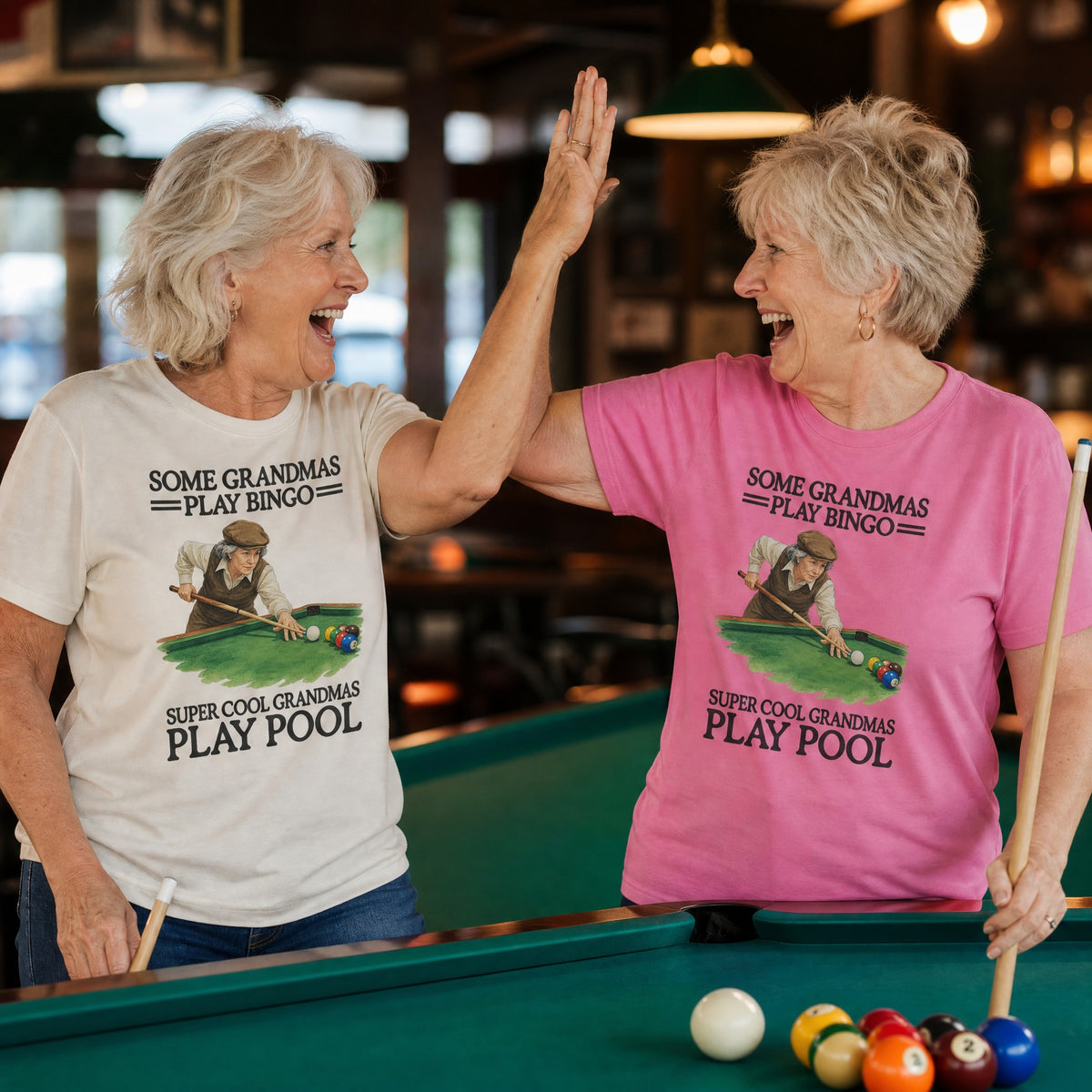 Two grandmas high-fiving at a pool table in the Super Cool Grandmas Play Pool tee, Ivory and Peony