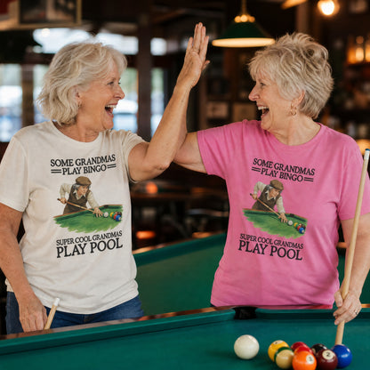 Two grandmas high-fiving at a pool table in the Super Cool Grandmas Play Pool tee, Ivory and Peony